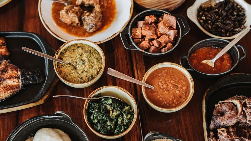 A spread of various Brazilian dishes including meat, beans, greens, and stews in bowls and plates on a wooden table.