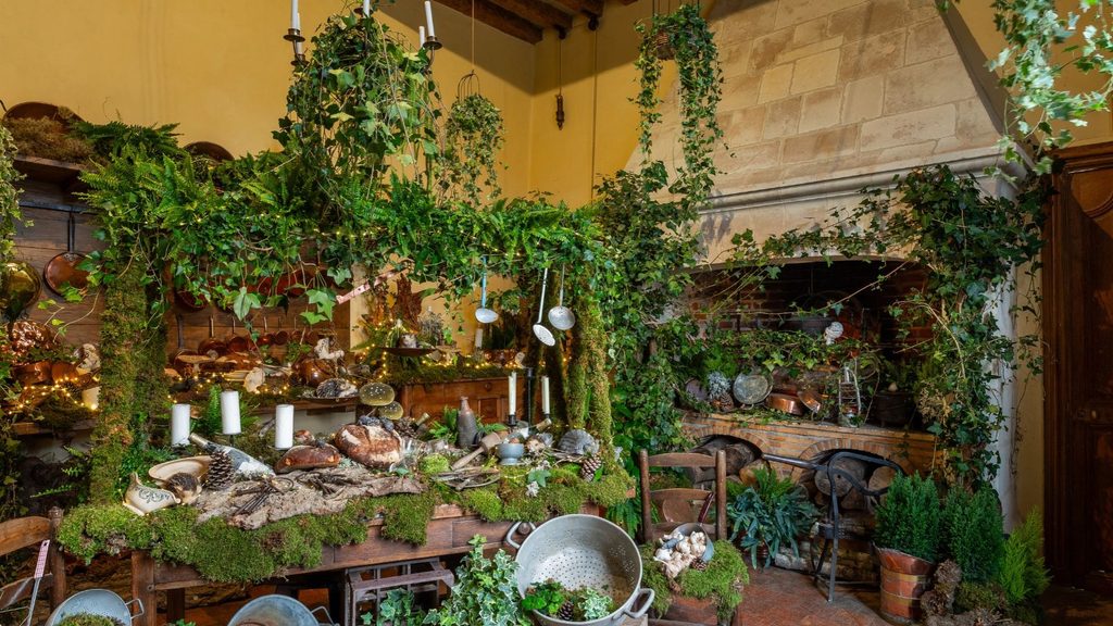 A rustic wooden table covered in moss, greenery, and fairy lights for a Christmas in the land of ch&acirc;teaux, featuring bread and kitchenware in a room with a stone fireplace.