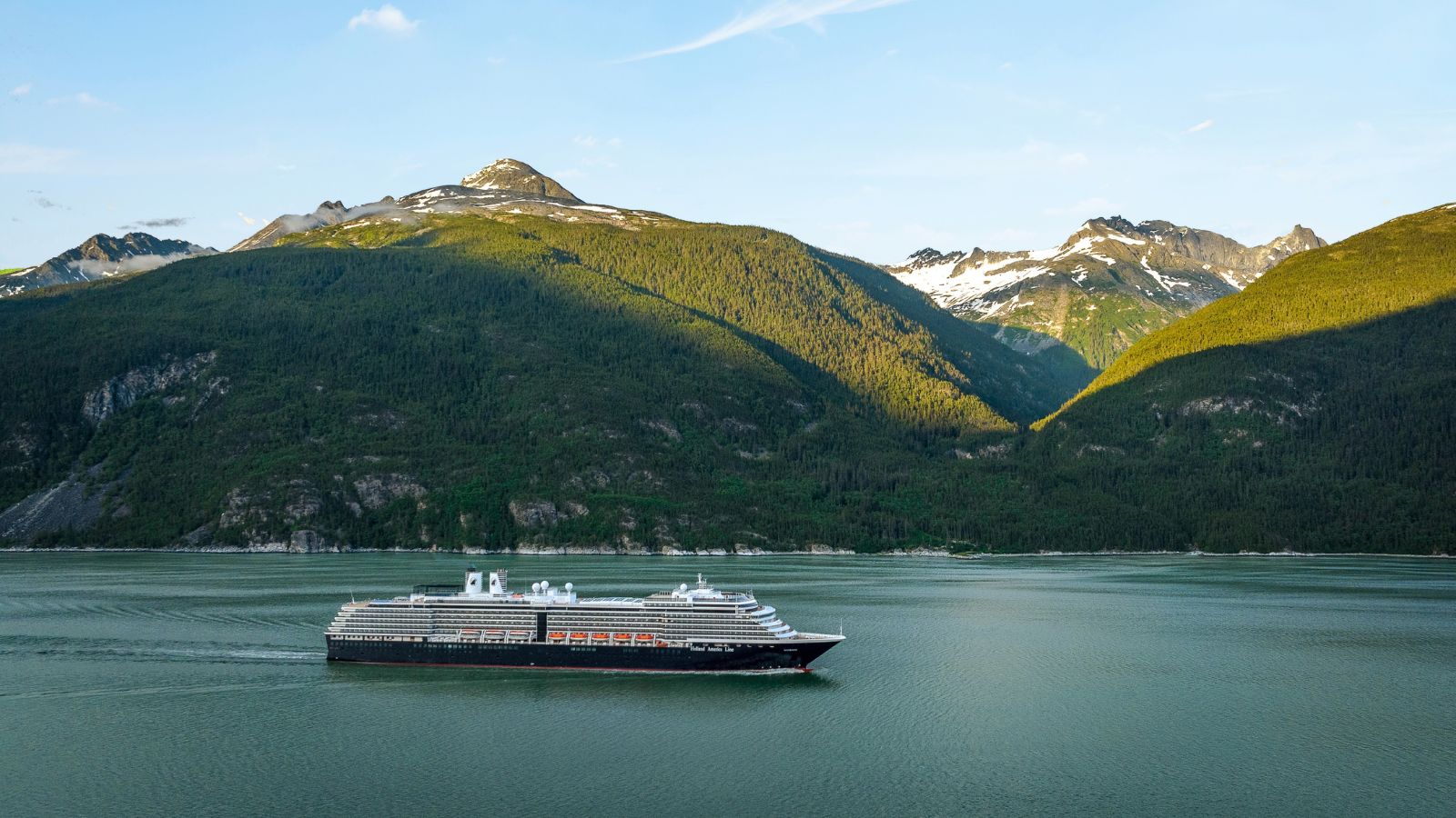 A Holland America Line cruise ship sailing through a narrow bay flanked by high, snow-capped mountains.