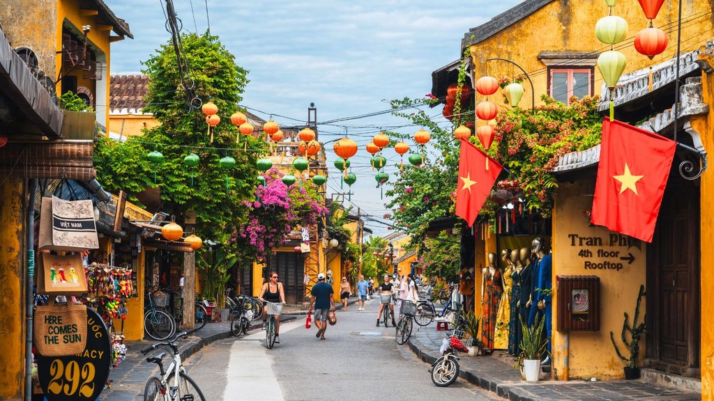 People walking and cycling down a narrow street in Hoi An, Vietnam, lined with yellow buildings and colorful paper lanterns.