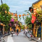People walking and cycling down a narrow street in Hoi An, Vietnam, lined with yellow buildings and colorful paper lanterns.