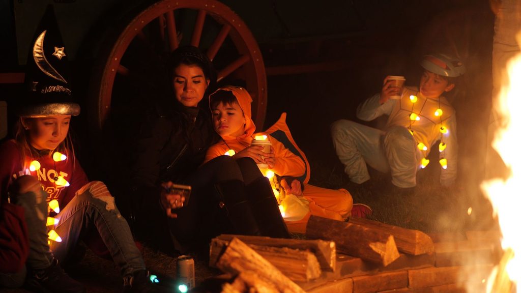 A family in Halloween costumes sits around a campfire at night.