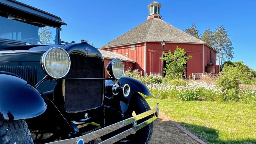 A black vintage Ford car parked in front of a red round barn.