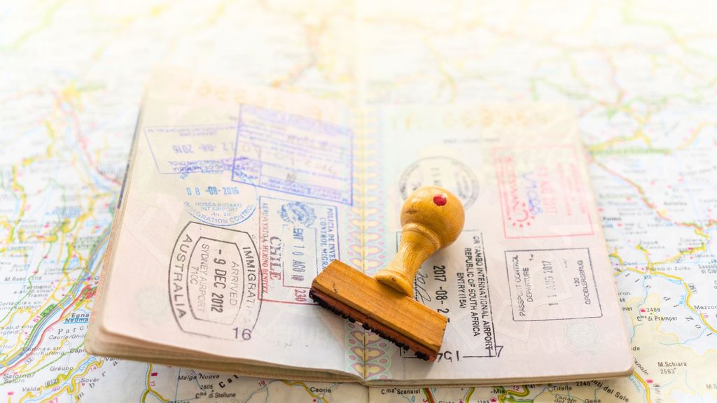 An open passport filled with Travelers Century Club international ink stamps, resting on a world map with a wooden rubber stamp on top.