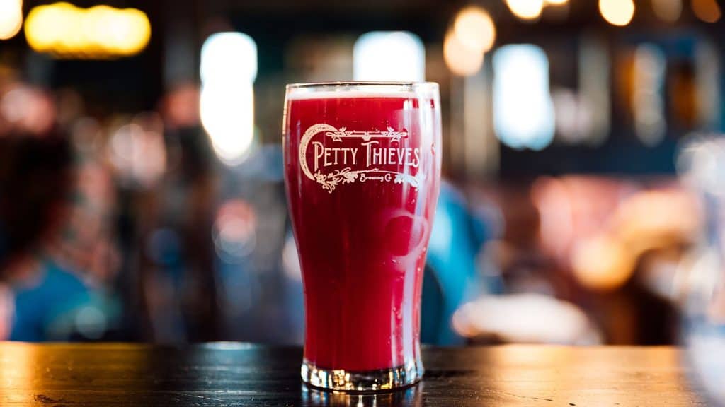 A pint glass of dark red beer featuring the Petty Thieves Brewing Co. logo, sitting on a wooden bar top.