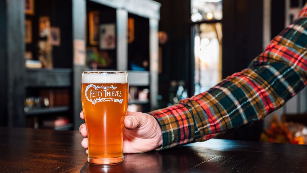 A person in a plaid flannel shirt holds a glass of beer with a “Petty Thieves” logo on a dark wooden bar at one of the Charlotte breweries.