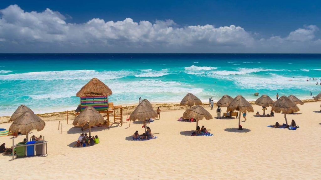 People relaxing on a sandy beach under thatched-roof umbrellas near a lifeguard tower and turquoise ocean water.