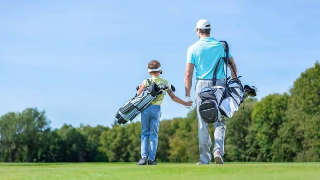 A man and a young child, both carrying golf bags, walk across a golf course.