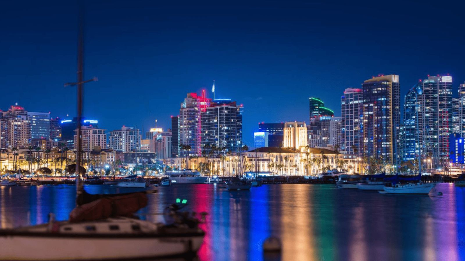 San Diego city skyline at night viewed from the harbor with boats in the foreground, glowing against the backdrop of light pollution.