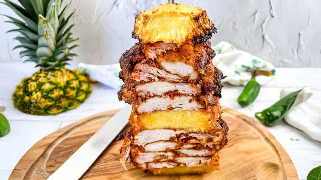 A vertical stack of sliced, seasoned pork and pineapple rings on a wooden cutting board, with a whole pineapple and peppers nearby.