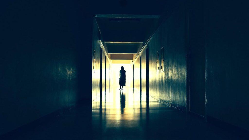 A silhouetted figure stands in the bright doorway at the end of a long, dark hallway.