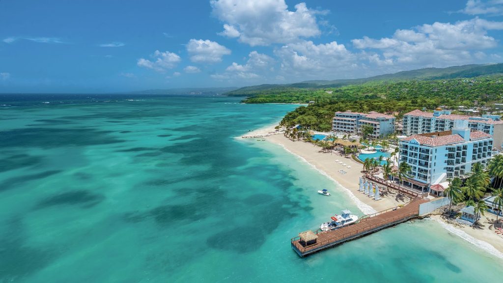 Aerial view of a Royal Caribbean Group coastal resort with large white buildings, swimming pools, a sandy beach, and a wooden pier extending into turquoise water.