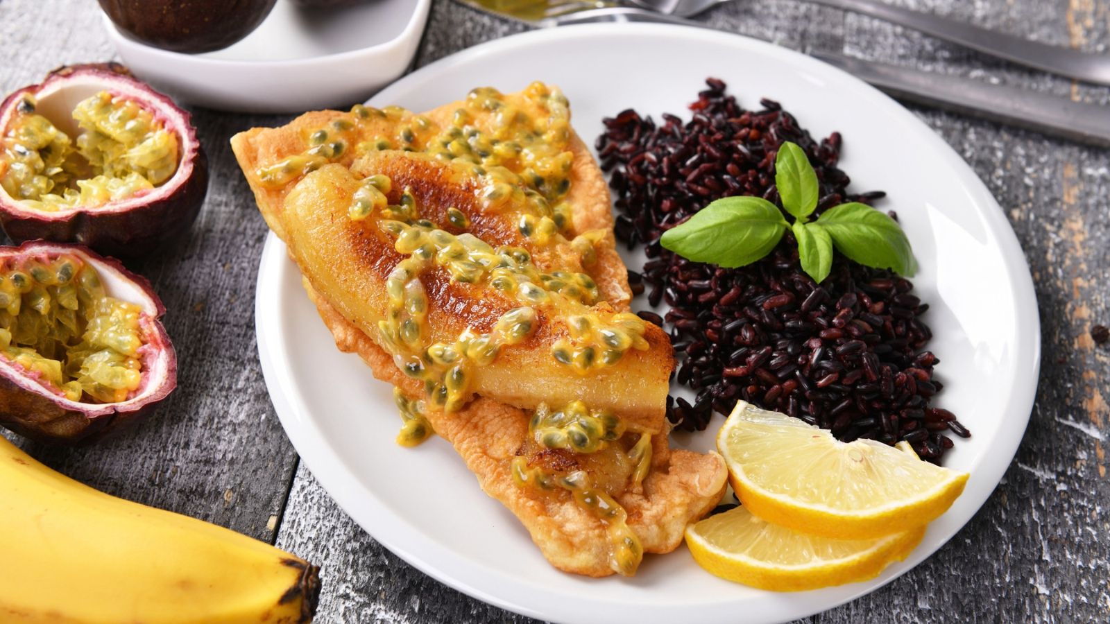 Fried fish topped with sliced banana and passion fruit sauce, served with black rice, lemon slices, and fresh basil.