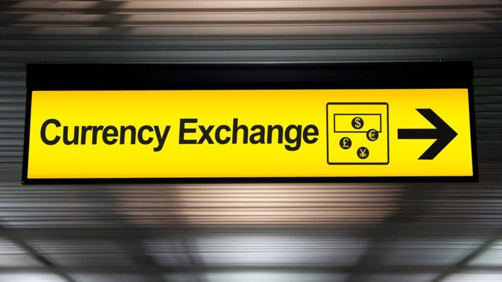 Yellow Currency Exchange sign with an icon of various currency symbols and a right-pointing arrow, marking common airport currency exchange traps.