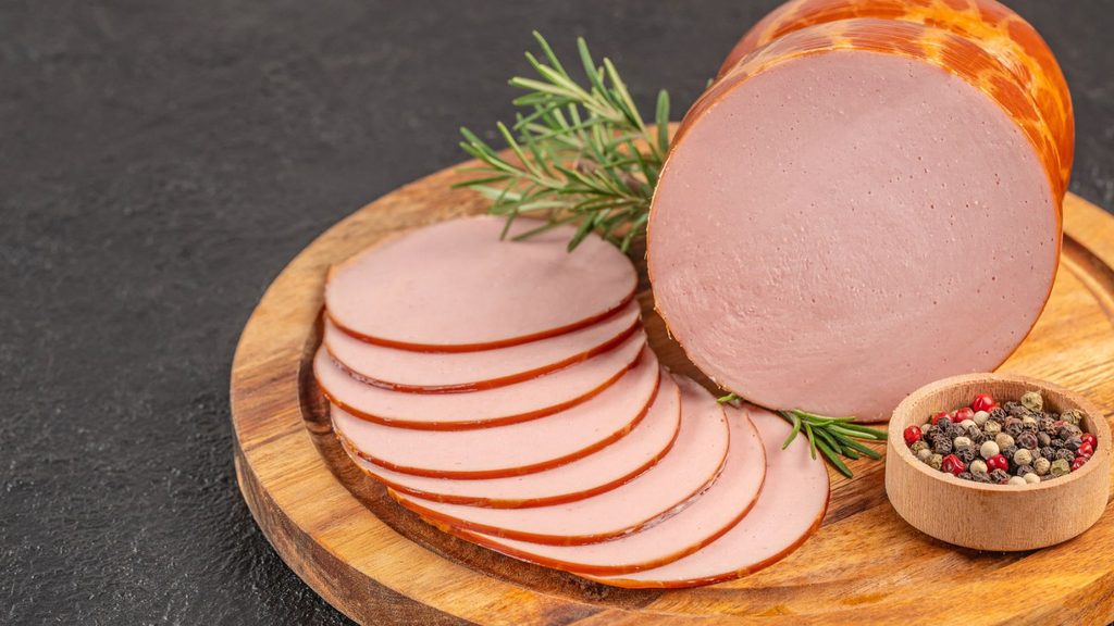A log of bologna and several slices arranged on a wooden board for National Bologna Day, garnished with rosemary and a small bowl of peppercorns.