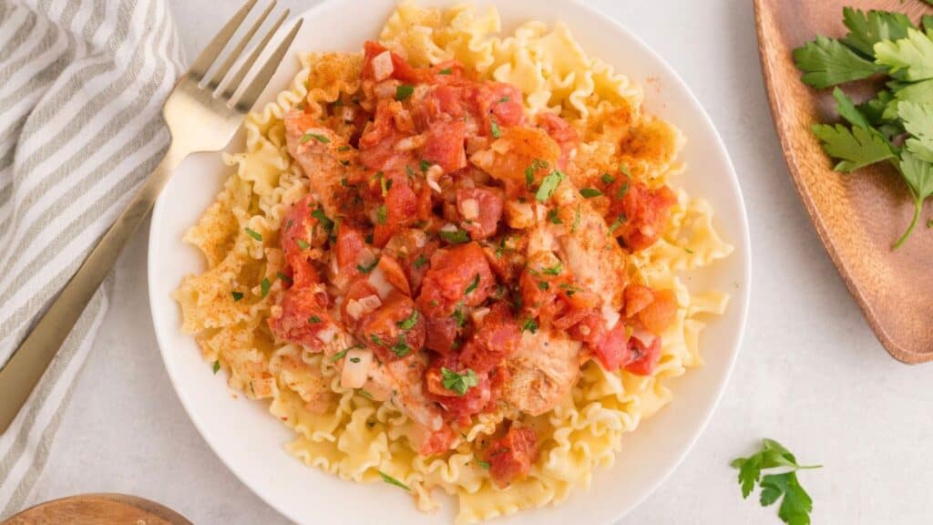 Chicken topped with diced tomatoes and herbs over curly pasta in a white bowl with a fork.