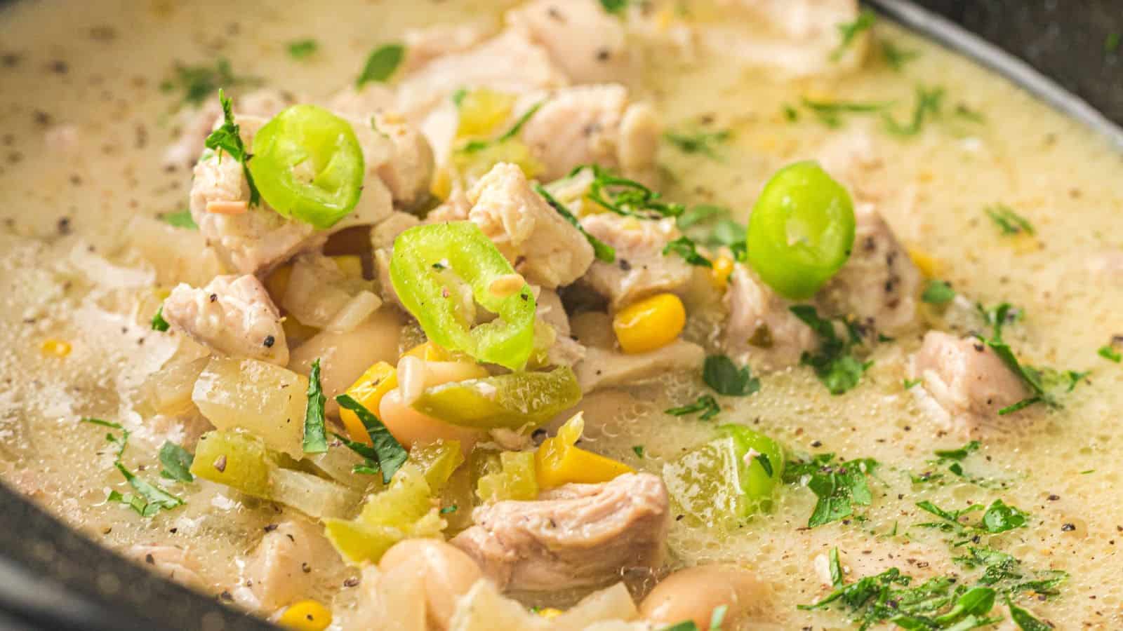 A close-up of white chicken chili containing shredded chicken, white beans, corn, and sliced green peppers.