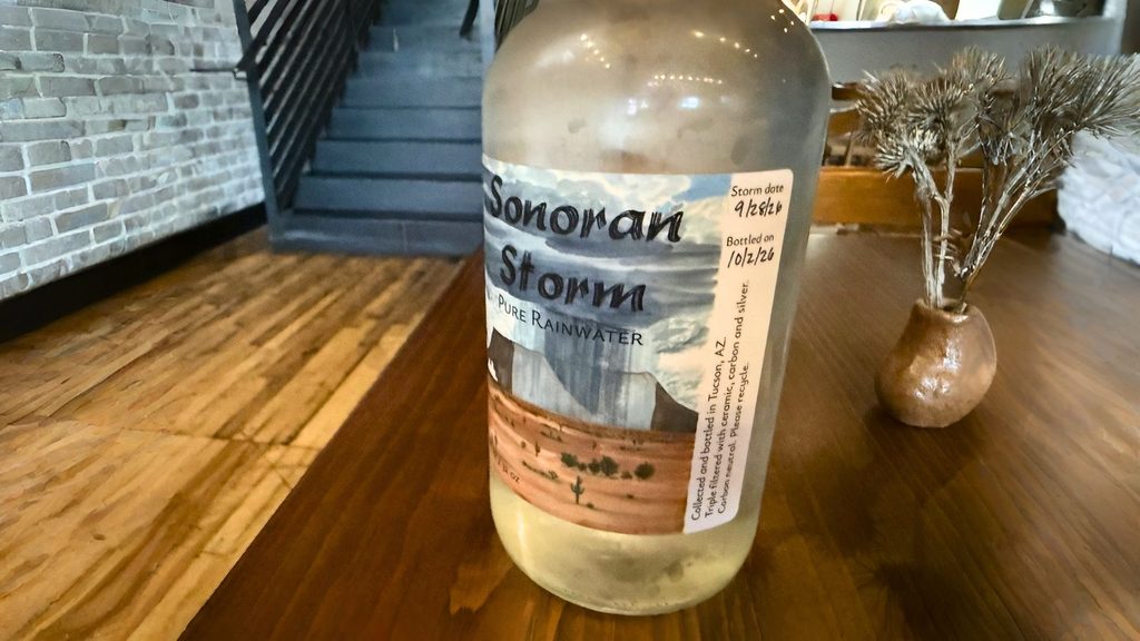 A glass bottle of Sonoran Storm Pure Rainwater from Tucson, with a sticker noting a storm date of 9/28/26 and a bottled date of 10/2/26.