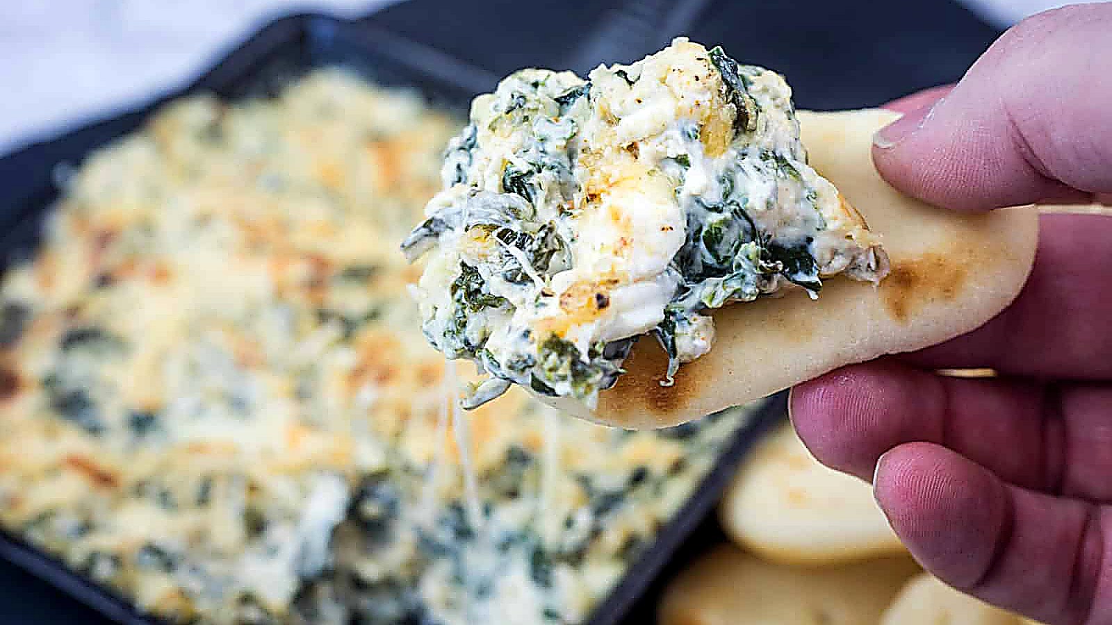 A hand holds a piece of flatbread topped with creamy spinach artichoke dip. A baking dish with more dip and a stack of flatbreads are in the background.