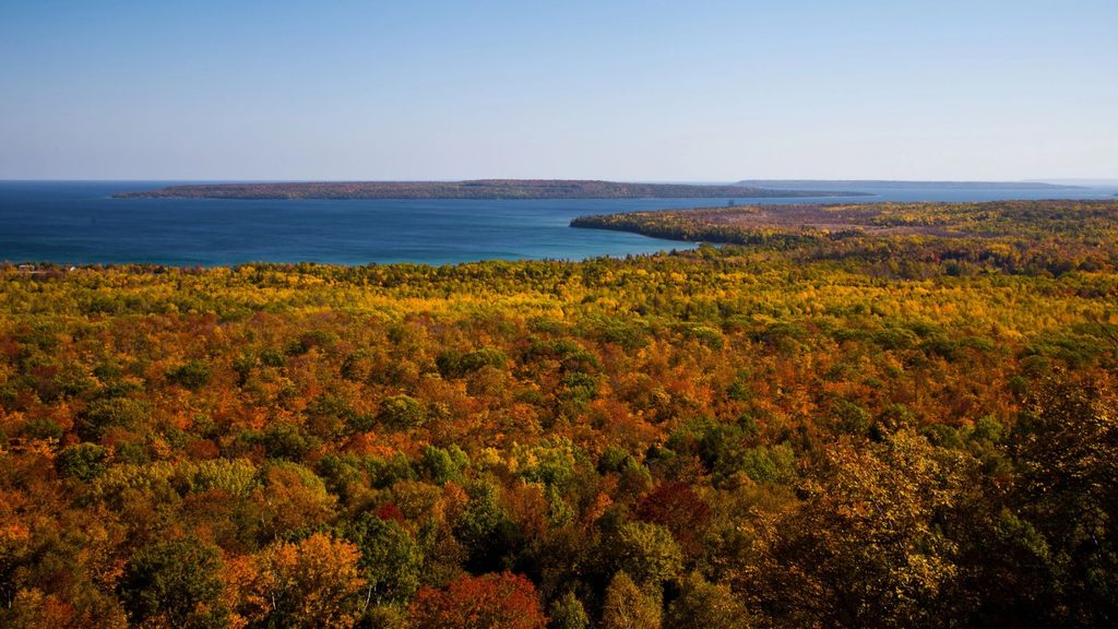 A vast forest canopy of autumn foliage in orange, yellow, and green overlooks a blue bay and distant peninsula, typical of Great Lakes harbor towns.