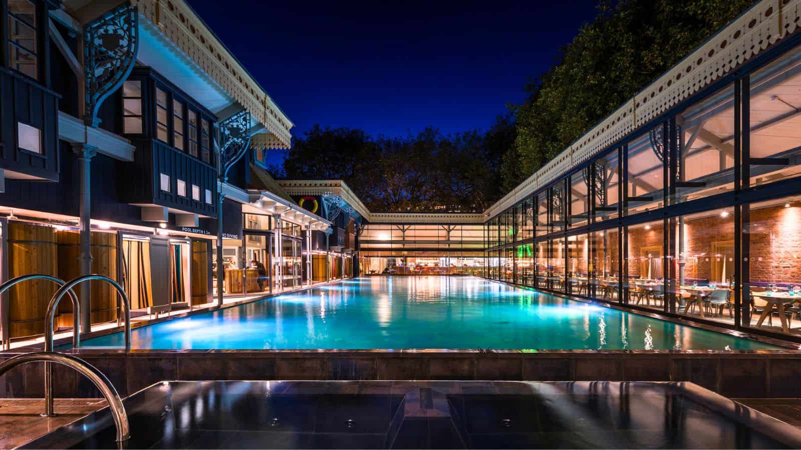 An illuminated Thames Lido swimming pool surrounded by dark buildings at night, with a glass-walled dining area on the right.