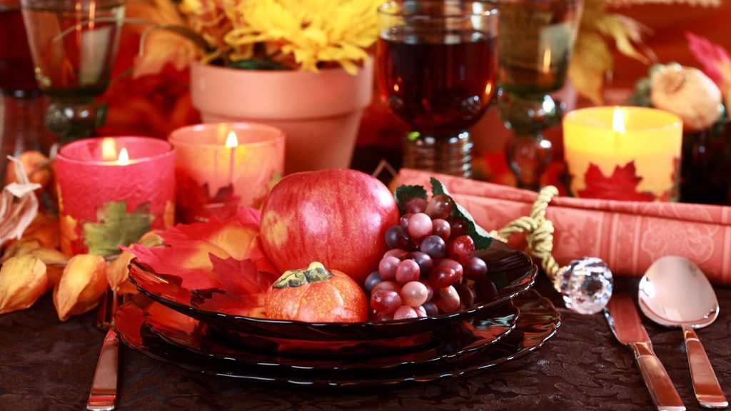 A dinner plate with an apple, a small gourd, and grapes, surrounded by Thanksgiving staples, candles, and glassware on a table.