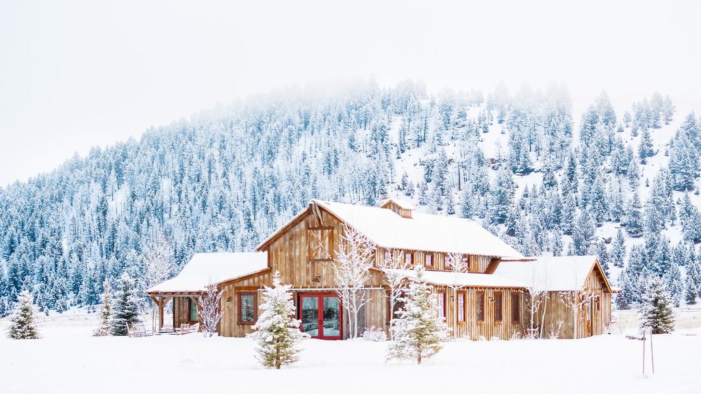 A wooden building with red doors sits in a snow-covered landscape with a pine forest in the background.