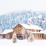 A wooden building with red doors sits in a snow-covered landscape with a pine forest in the background.