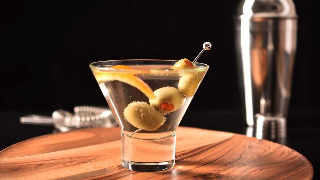 A tiny cocktail martini in a stemmed glass garnished with three olives on a pick and a citrus twist, sitting on a wooden board with a cocktail shaker in the background.