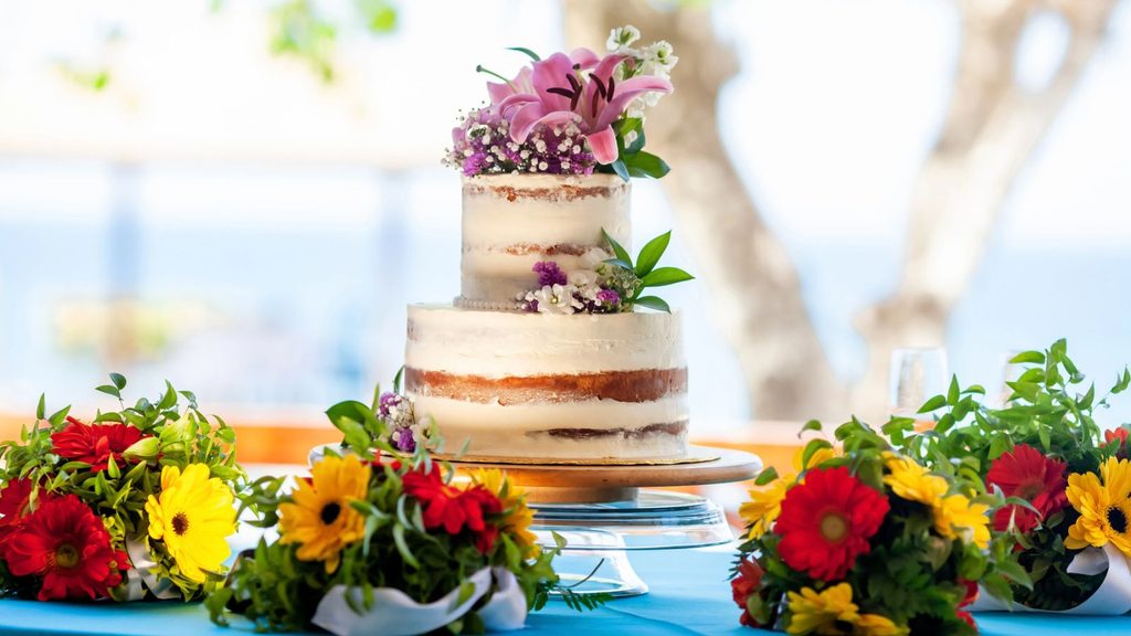 Two-tier semi-naked wedding cake decorated with pink lilies and white flowers, centered on a table with bouquets of red and yellow gerbera daisies.