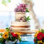 Two-tier semi-naked wedding cake decorated with pink lilies and white flowers, centered on a table with bouquets of red and yellow gerbera daisies.