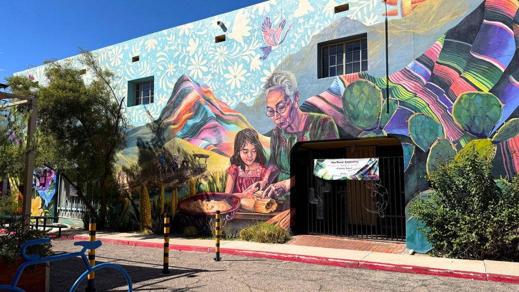 Large mural on a building exterior depicting an older woman and a young girl preparing food, set against a backdrop of colorful mountains and desert plants.