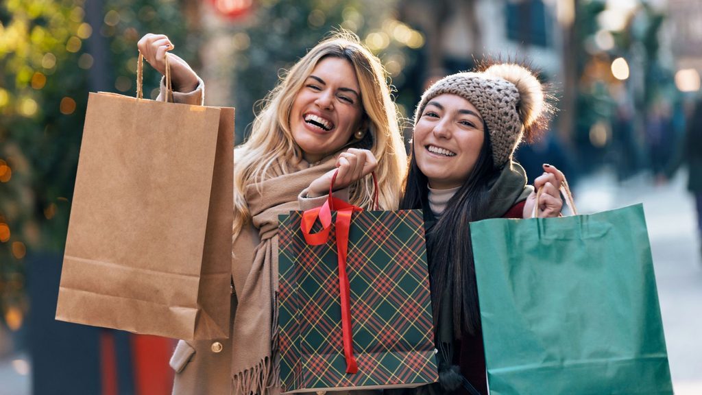 Two women smiling and holding bags while out early holiday shopping.