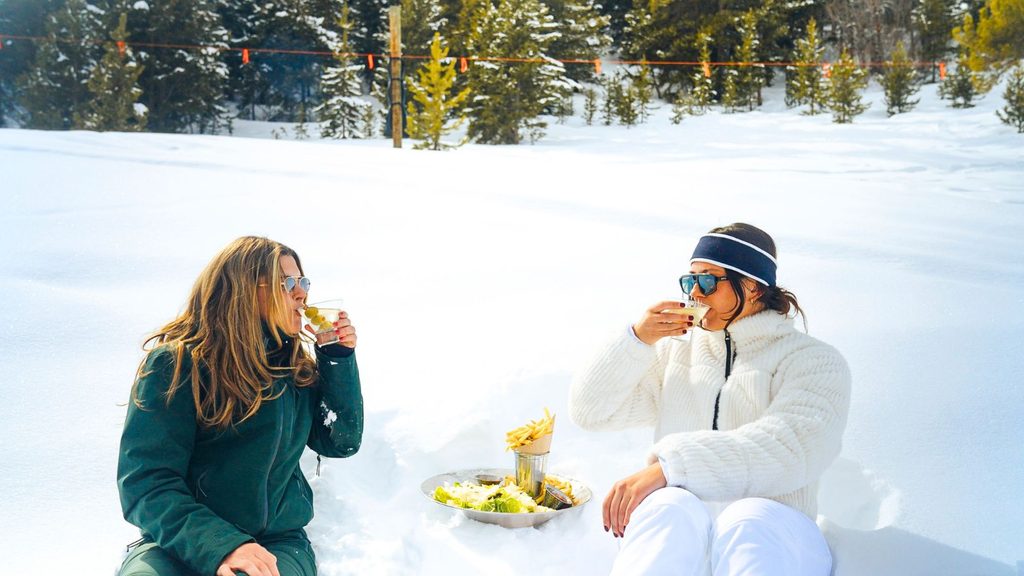 Two women in winter clothing sit in the snow, drinking cocktails and eating fries from a platter.