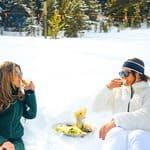 Two women in winter clothing sit in the snow, drinking cocktails and eating fries from a platter.