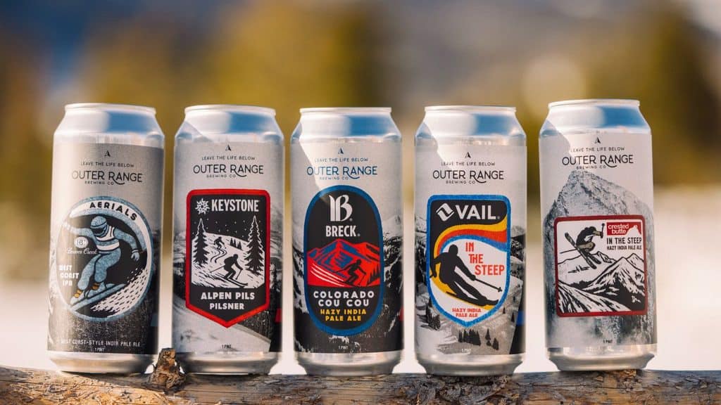 Five cans of Outer Range Brewing Co. beer lined up on a wooden surface, each featuring unique labels with skiing and mountain-themed graphics.