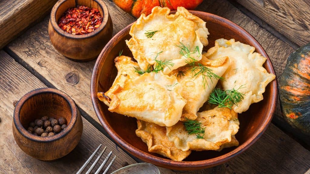 A bowl of fried pierogi garnished with dill for National Pierogi Day, next to small bowls of red pepper flakes and peppercorns.
